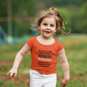 little girl wearing an orange shirt that says halloweentown correctional tresspassing walked like a zombie in a graveyard