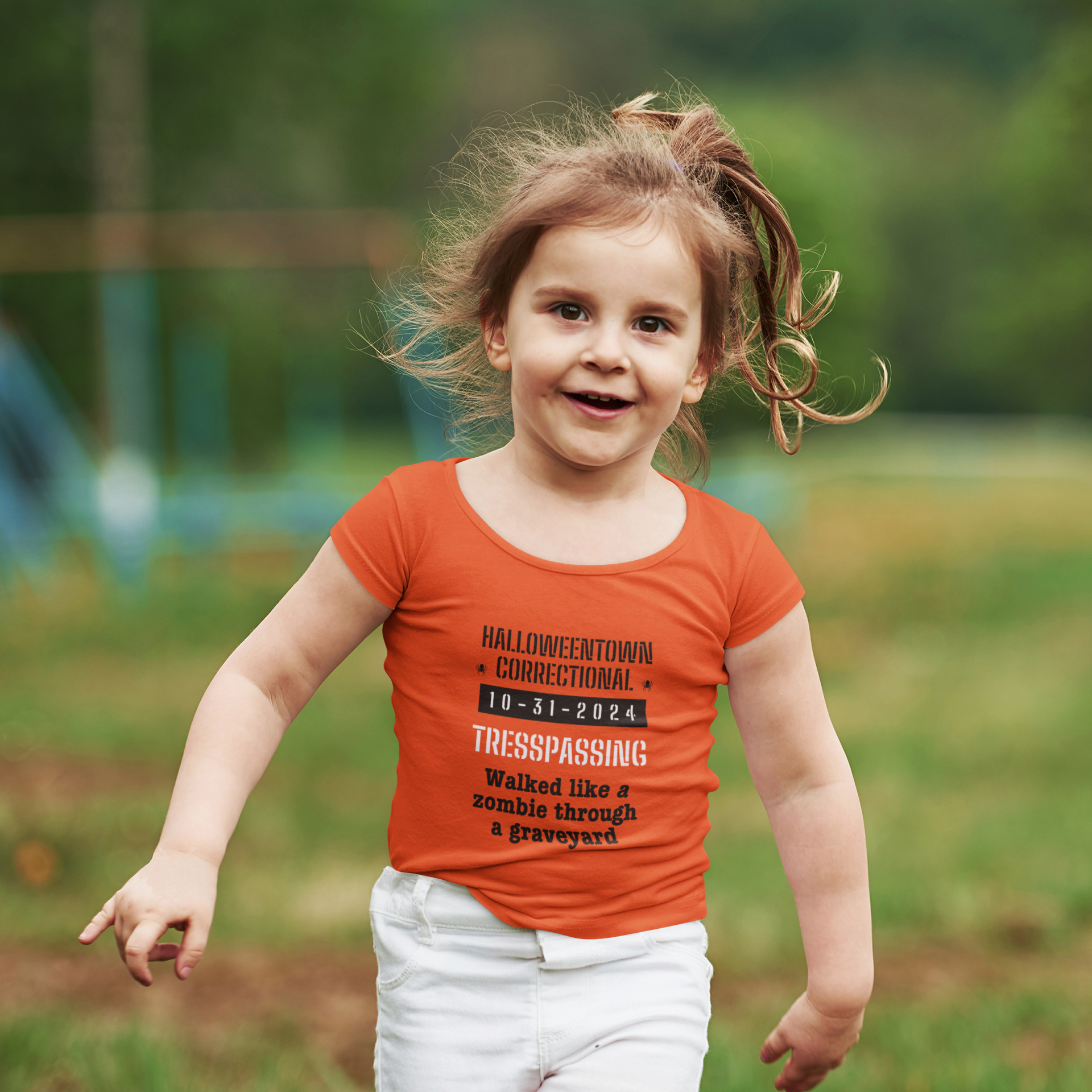 little girl wearing an orange shirt that says halloweentown correctional tresspassing walked like a zombie in a graveyard