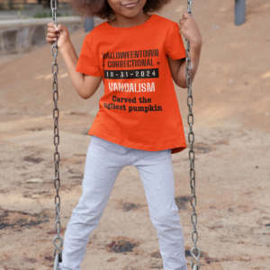 kid on swing wearing orange shirt that says halloweentown correctional vandalism carved the ugliest pumpkin