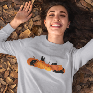 woman laying on leaves wearing a halloween sweatshirt with black cats and pumpkins